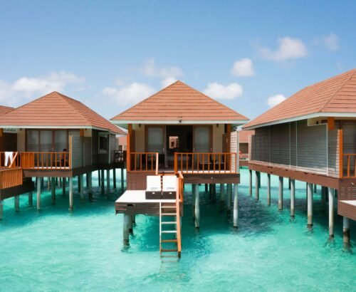 Deluxe Water Villas at Brennia Kottefaru Maldives standing on stilts above the clear turquoise lagoon with wooden decks and stairs leading into the water under a bright blue sky