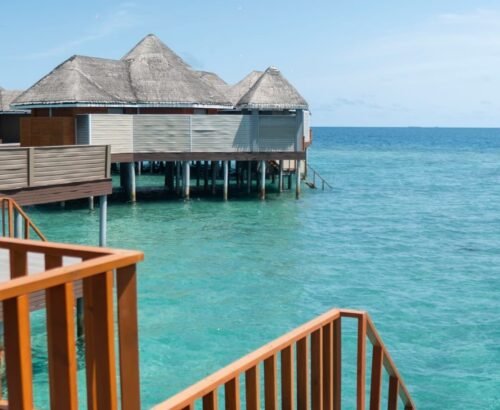 Deluxe Water Villas at Brennia Kottefaru Maldives with thatched roofs standing on stilts above the clear turquoise lagoon, viewed from a wooden deck under a bright blue sky