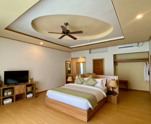 Spacious Deluxe Water Villa bedroom at Brennia Kottefaru Maldives with a large wooden bed, TV console, open wardrobe with bathrobes, and warm natural lighting