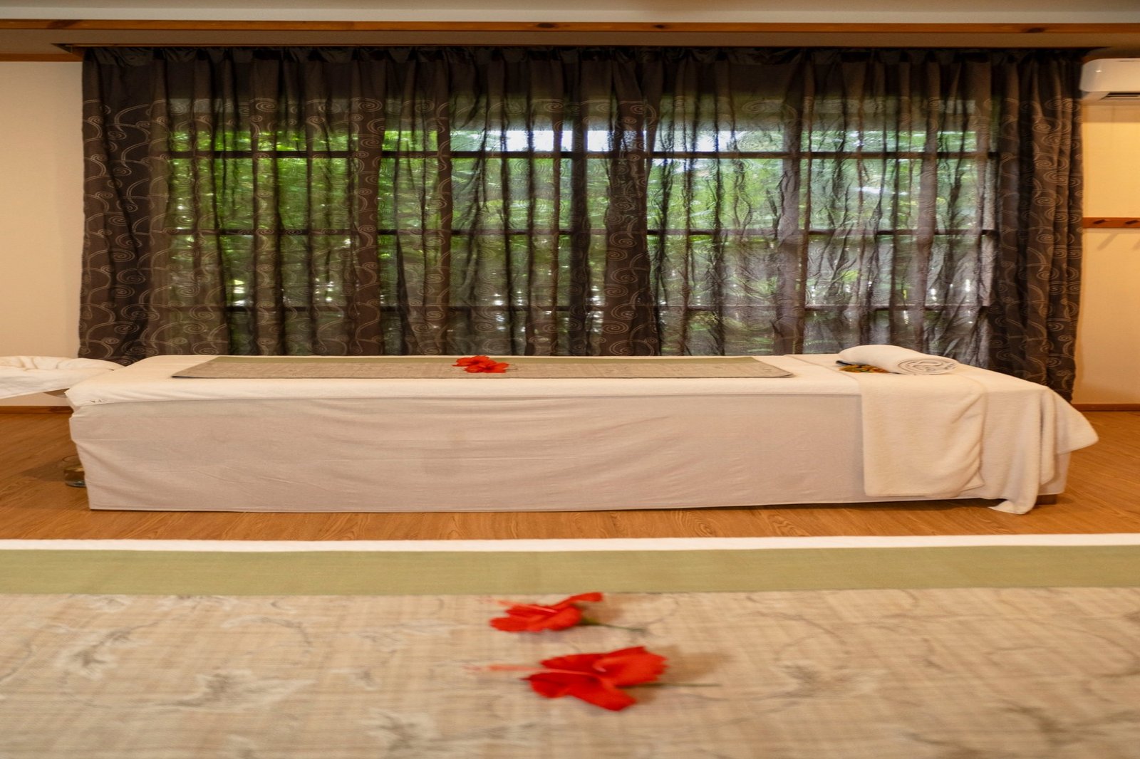 Peaceful spa treatment room at Brennia Kottefaru Maldives featuring a massage bed with soft linens, red flower petals, and large windows overlooking tropical greenery