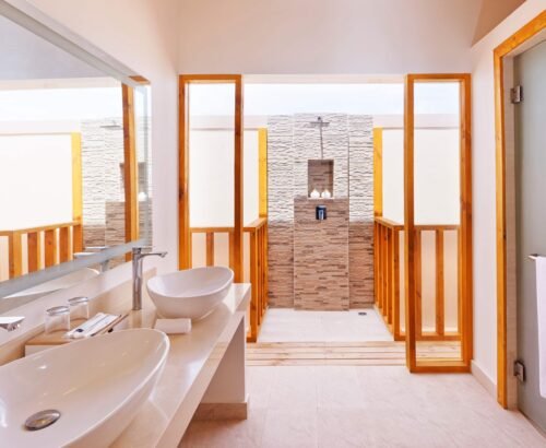 Ocean Pool Villa bathroom at Brennia Kottefaru Maldives featuring dual white vessel sinks on a long vanity and an open stone-tiled outdoor shower framed by wooden panels