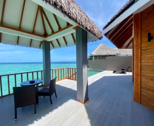 Covered wooden deck of an Over Water Suite with Pool at Brennia Kottefaru Maldives featuring outdoor seating, a private pool, and open views of the turquoise lagoon under a clear blue sky