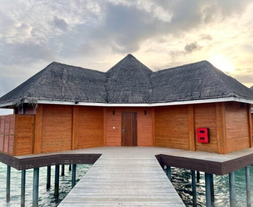 Exterior of an Over Water Suite with Pool at Brennia Kottefaru Maldives featuring wooden walls, thatched roof, and a wooden walkway leading to the entrance over the lagoon at sunset