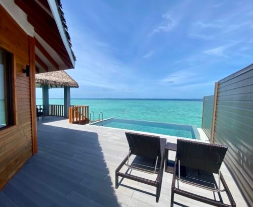 Private wooden deck of an Over Water Suite with Pool at Brennia Kottefaru Maldives featuring sun loungers, an infinity-style pool, and open views of the turquoise lagoon under a clear blue sky