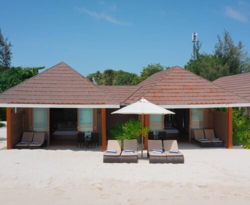 Two Bedroom Beach Villas at Brennia Kottefaru Maldives with open bedrooms facing the white sand beach, sun loungers, and white umbrellas under a clear blue sky