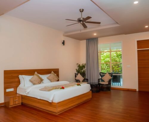Spacious bedroom in a Two Bedroom Bougain Villa at Brennia Kottefaru Maldives featuring a large wooden bed with floral décor, seating area by the window, and views of tropical greenery outside