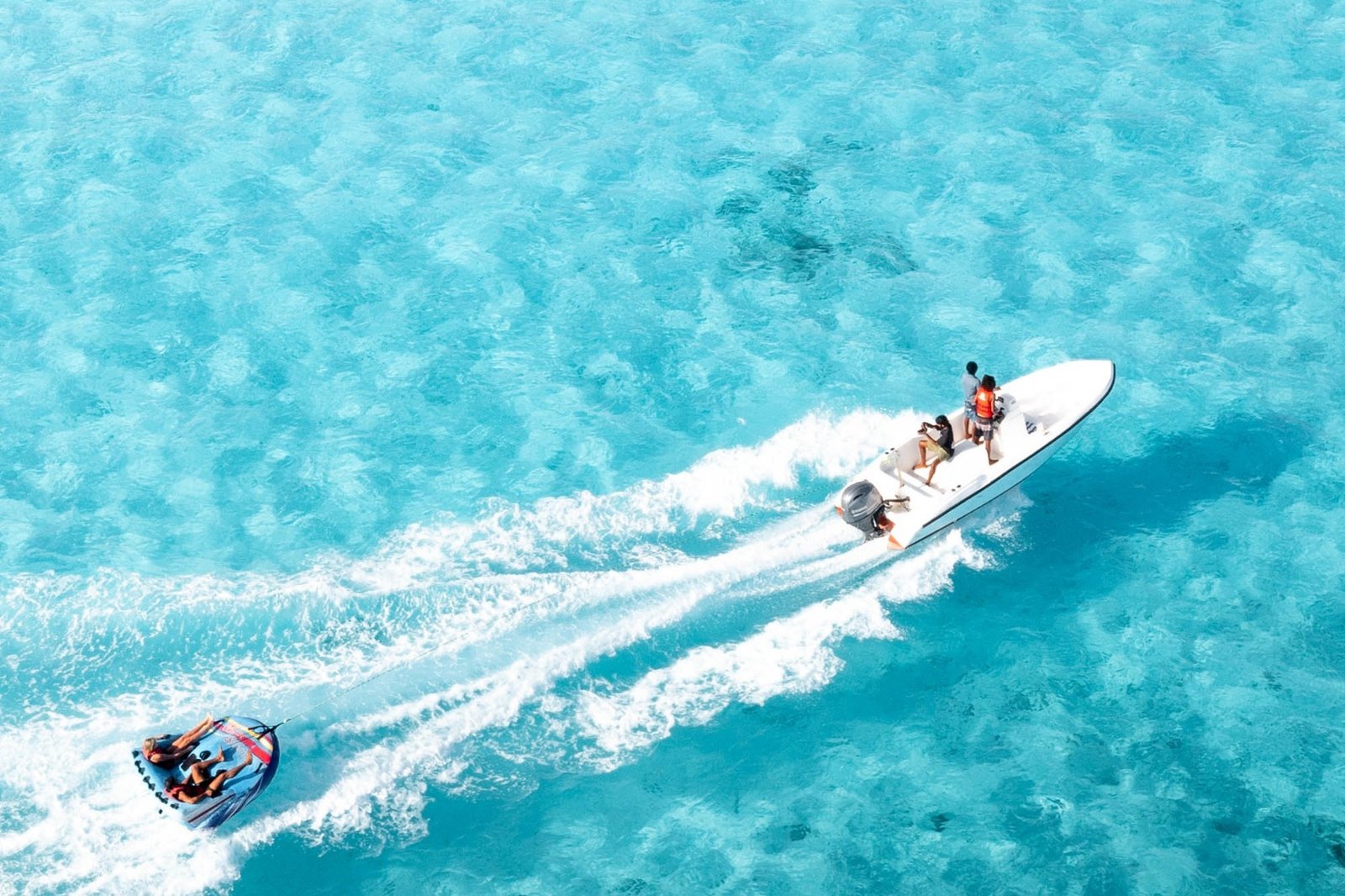 Aerial view of water sports at Brennia Kottefaru Maldives showing a speedboat towing a water tube with guests across the clear turquoise lagoon