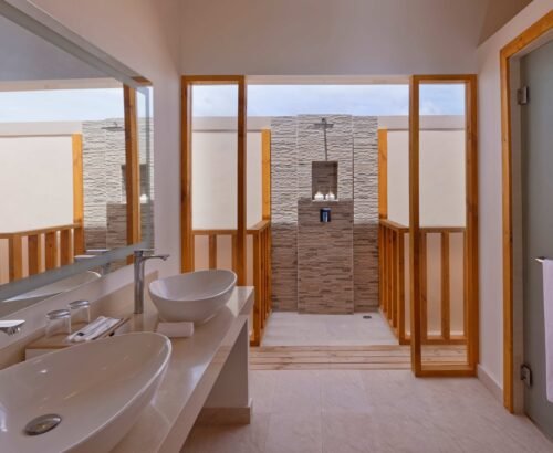 Water Villa bathroom at Brennia Kottefaru Maldives featuring dual white vessel sinks on a long vanity and an open stone-tiled outdoor shower framed by wooden panels