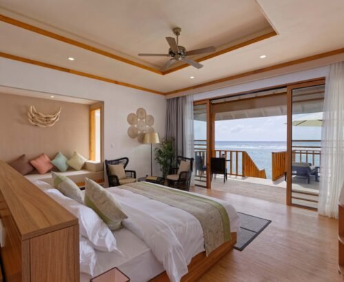 Spacious Water Villa bedroom at Brennia Kottefaru Maldives with a large bed, built-in daybed, seating area, and open sliding doors leading to an overwater deck with ocean views