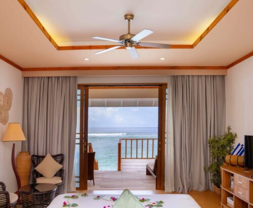 Water Villa bedroom at Brennia Kottefaru Maldives with a bed decorated with flowers, seating area, and open sliding doors leading to a wooden deck overlooking the turquoise ocean
