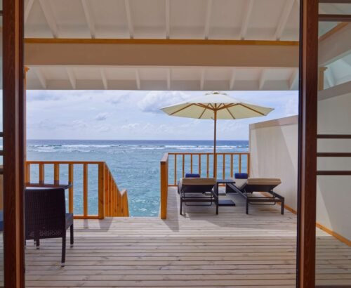 Private wooden deck of a Water Villa at Brennia Kottefaru Maldives with sun loungers, white umbrella, and open views of the turquoise ocean under a blue sky