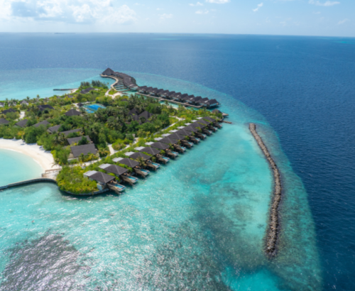 Aerial view of dusitD2 Feydhoo Resort Maldives showing waterfront pool villas extending over the crystal-clear turquoise lagoon, lush tropical island, white sand beach, and deep blue ocean under a bright sky – the perfect setting for a Maldives tour package.