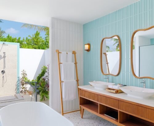 Luxurious bathroom in the beach pool villa at dusitD2 Feydhoo Resort Maldives featuring dual vessel sinks, wooden vanity, freestanding bathtub, and open-air outdoor shower surrounded by tropical plants under a blue sky – perfect for a Maldives tour package experience.