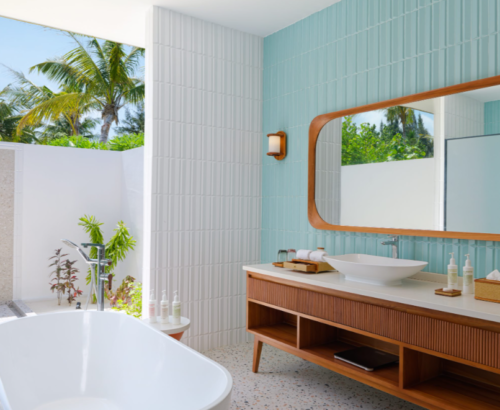 Elegant bathroom in the beach villa at dusitD2 Feydhoo Resort Maldives featuring a freestanding bathtub, outdoor shower with tropical plants and palm trees, wooden vanity with vessel sink, and large wooden-framed mirror – ideal for a Maldives tour package stay.