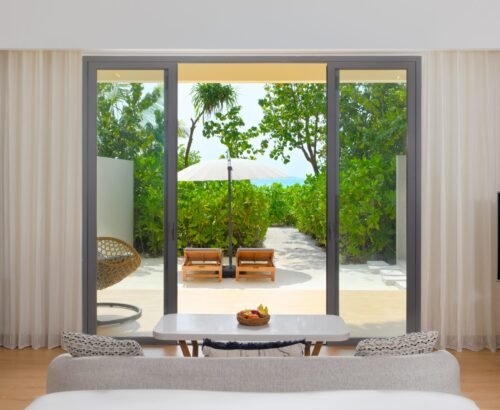 Bright beach villa bedroom interior at dusitD2 Feydhoo Resort Maldives with large glass doors opening to a private outdoor deck, sun loungers, hanging chair, tropical garden, white sand path, and distant ocean view – ideal for a Maldives tour package stay.