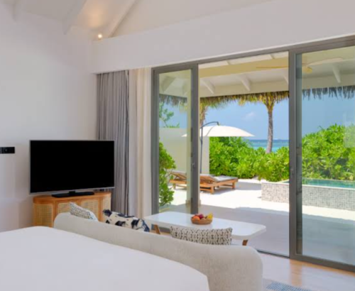 Bright beach villa bedroom at dusitD2 Feydhoo Resort Maldives with large glass doors opening to a private outdoor deck, sun loungers, infinity pool, tropical greenery, and ocean views under a clear blue sky – ideal for a Maldives tour package stay.