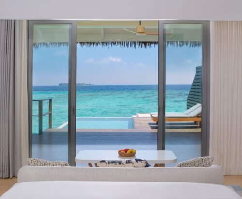 Luxurious bedroom interior at dusitD2 Feydhoo Resort Maldives with floor-to-ceiling glass doors opening to a private infinity pool, wooden deck, sun loungers, and turquoise lagoon ocean view – perfect for a Maldives tour package experience.