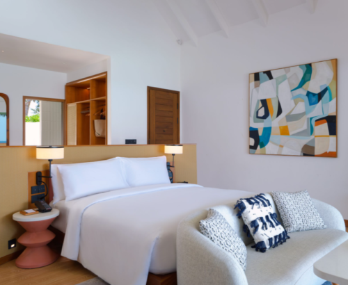 Stylish beach villa bedroom at dusitD2 Feydhoo Resort Maldives featuring a king-size bed with cane headboard, curved sofa, abstract wall art, open wardrobe, and a glimpse of the ocean through the doorway – perfect for a Maldives tour package stay.