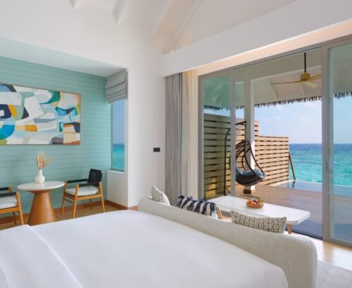 Bright overwater pool villa bedroom at dusitD2 Feydhoo Resort Maldives featuring a king-size bed, modern seating area, abstract wall art, large glass doors opening to a private wooden deck with hanging chair, infinity pool, and turquoise ocean views – perfect for a Maldives tour package.
