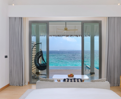 Bright overwater villa bedroom at dusitD2 Feydhoo Resort Maldives with large glass doors opening to a private wooden deck, hanging chair, infinity pool, and panoramic turquoise ocean views under a clear blue sky – ideal for a Maldives tour package.