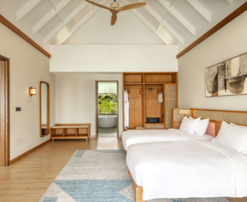 Spacious twin bedroom in the Two Bedroom Party Suite at dusitD2 Feydhoo Resort Maldives featuring two single beds with cane headboards, high vaulted ceiling, open wooden wardrobe, abstract wall art, and a view into the bathroom with freestanding bathtub – ideal for a Maldives tour package stay.